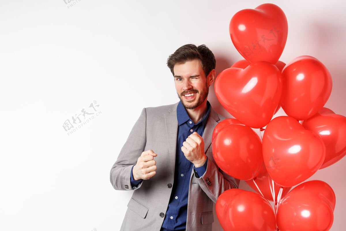 男朋友帅哥在情人节的心形气球旁翩翩起舞 微笑着 对着镜头眨眼 在情人节的浪漫约会中穿上衣服 白色背景浪漫气球微笑