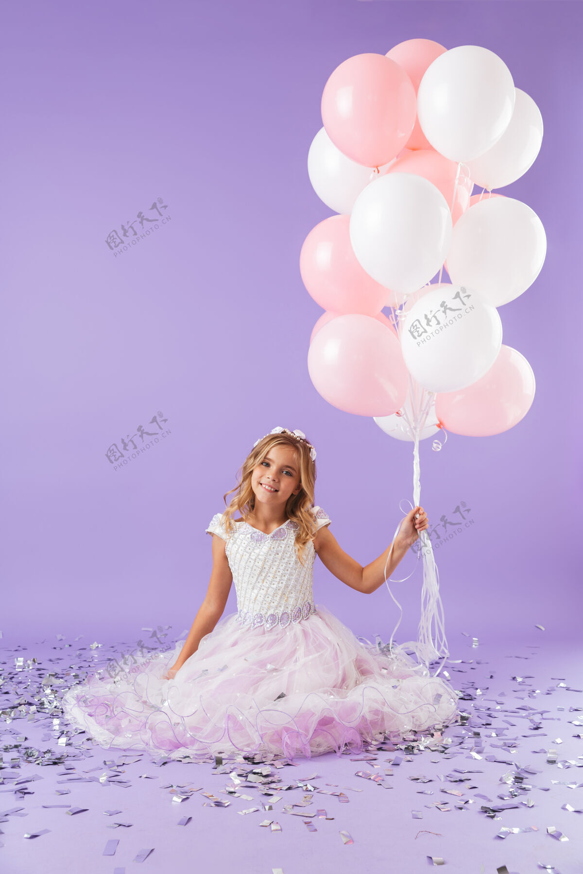 公主穿着公主裙的漂亮小女孩孤立地坐在紫罗兰色的墙上 手里拿着一堆气球小束童年