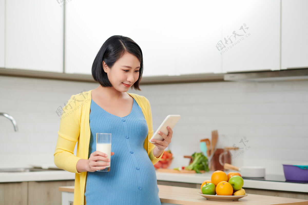 蛋白质年轻的孕妇拿着一杯牛奶在餐桌旁 厨房里有食物期望父母亚洲人