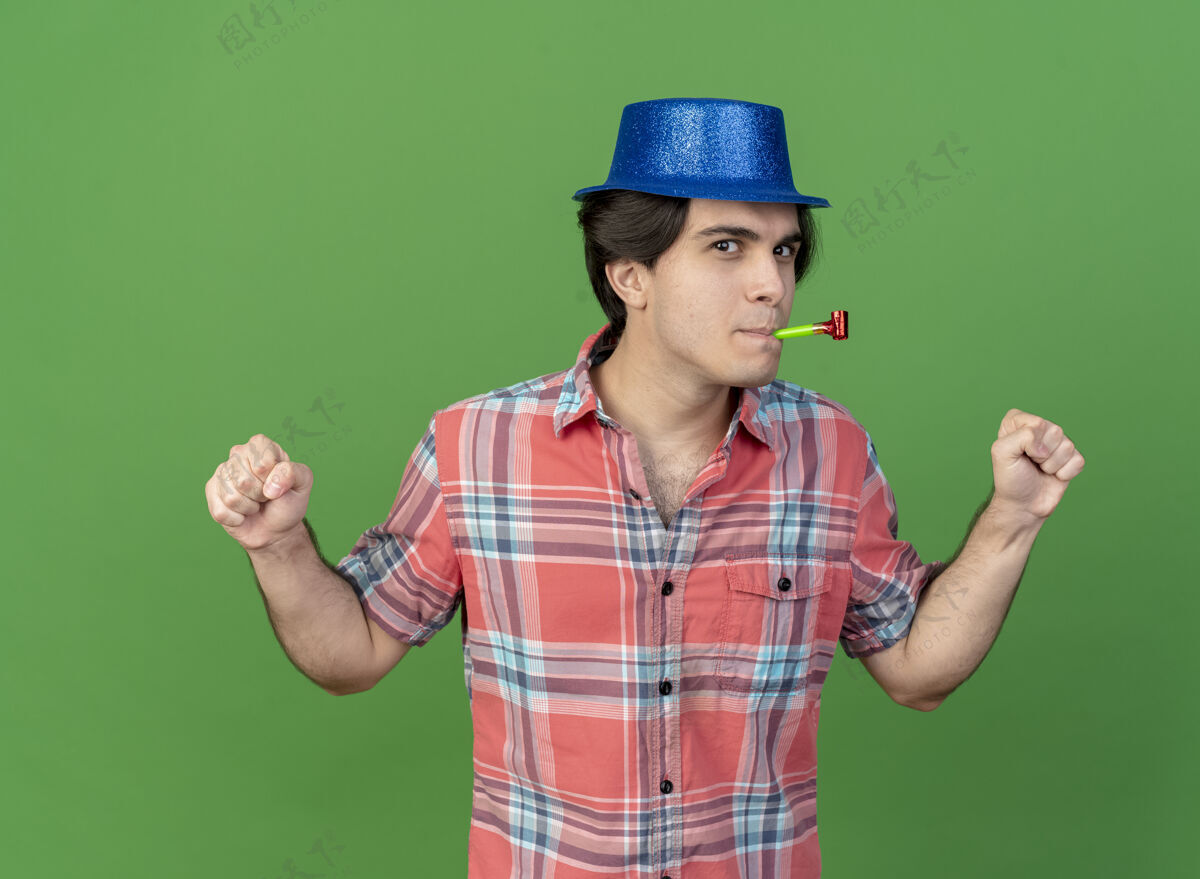 帽子戴着蓝色派对帽的白种人帅哥高兴地用拳头吹着派对哨子男人绿色请