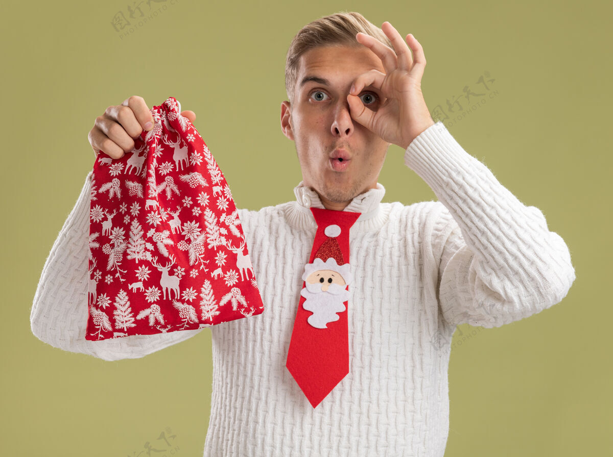 领带印象深刻的年轻帅哥戴着圣诞老人领带拿着圣诞袋看着相机做着看起来手势孤立在橄榄绿的背景上印象圣诞老人年轻