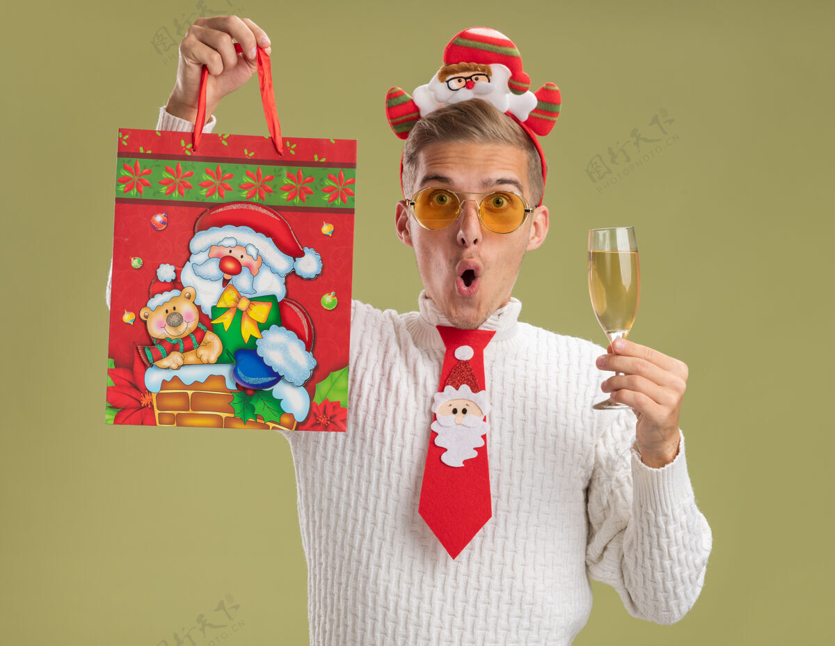 新年印象深刻的年轻帅哥戴着圣诞老人的头带和领带看着相机拿着一杯香槟和提高圣诞礼品袋孤立在橄榄绿的背景领带杯子礼物
