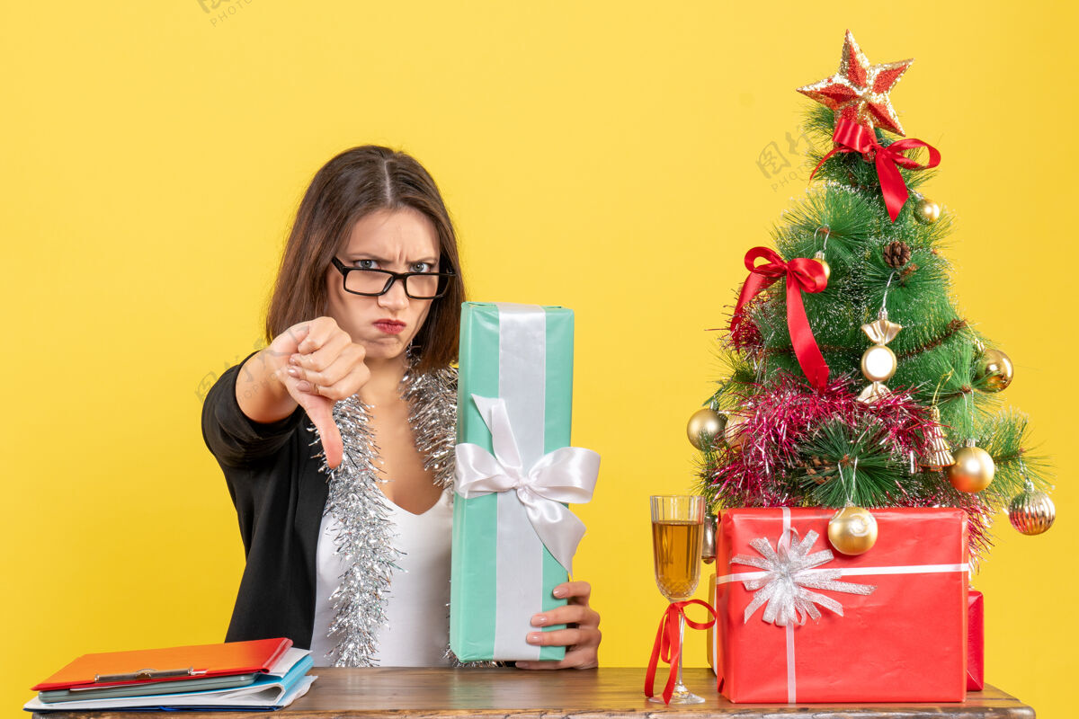 展示一位穿着西装戴着眼镜的漂亮女士在办公室里摆着一张桌子 桌上放着一棵圣诞树成人礼物手势