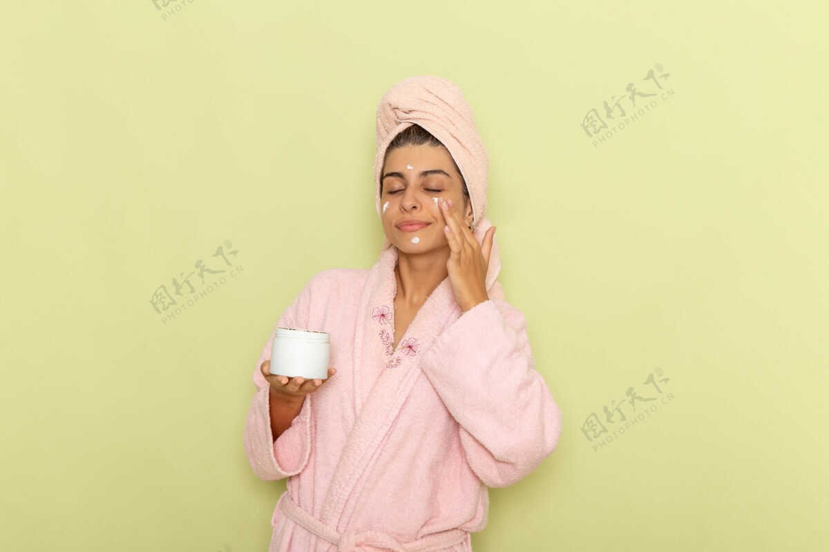 口罩正面图穿着粉色浴袍的年轻女性在绿色表面涂面霜自我护理浴衣乳霜