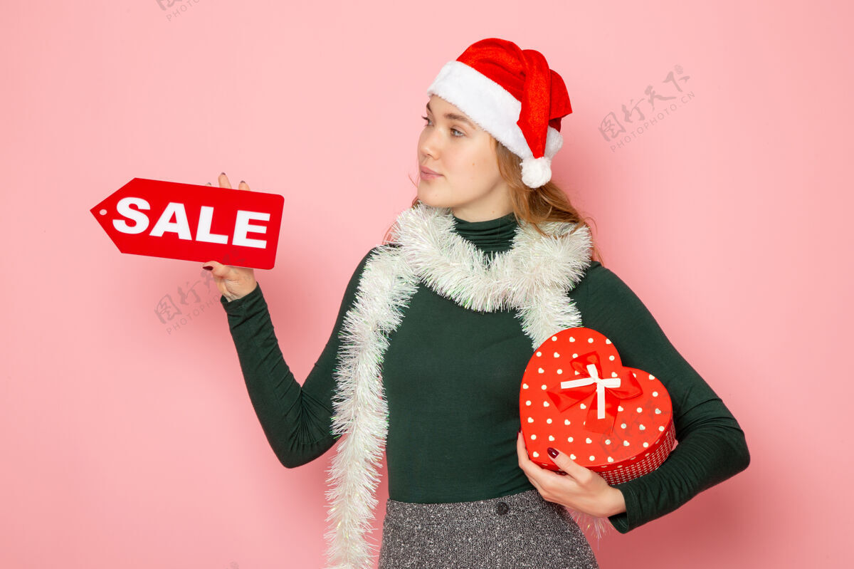 写作正面图年轻女子手捧红色大卖场 在粉色墙上书写和呈现圣诞新年购物情感节日色彩帽子肖像孩子