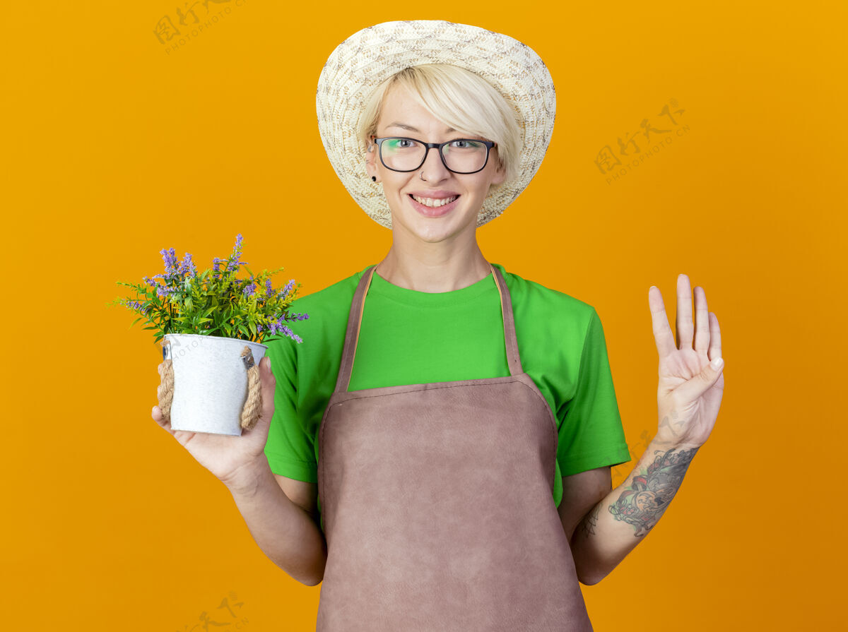年轻围裙上留着短发 戴着帽子 手里拿着盆栽植物的年轻园丁看着摄像机 脸上洋溢着幸福的笑容 四号站在橙色的背景上相机数字快乐