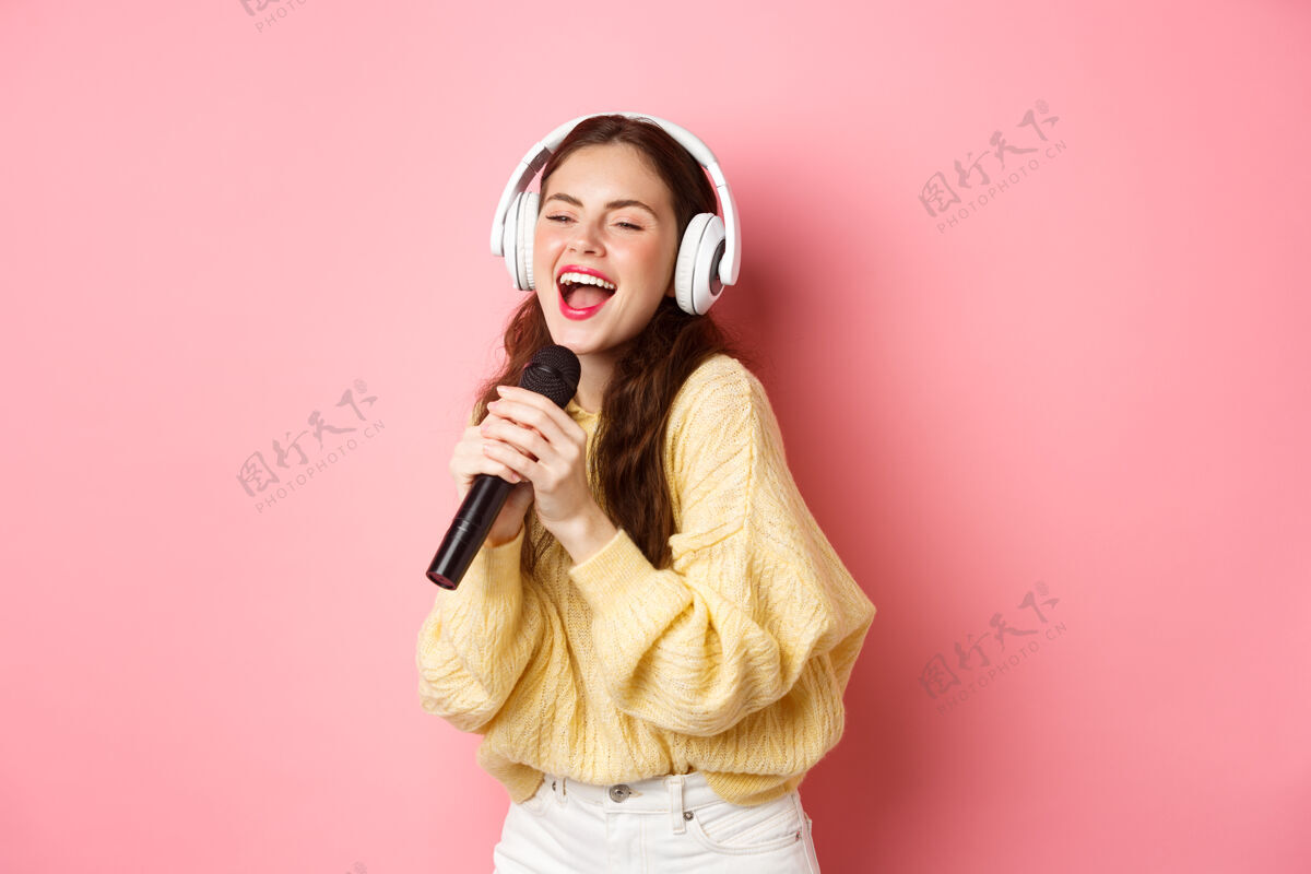 兴奋无忧无虑的女孩享受卡拉OK之夜 在麦克风里唱歌 戴着无线耳机 站在粉红色的墙上卡拉OK欢呼女孩