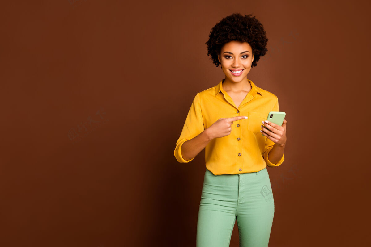 情绪正面开朗的黑人女学生肖像使用智能手机搜索网上销售指示点食指装置穿时尚黄色裤子隔离棕色微笑女人美国