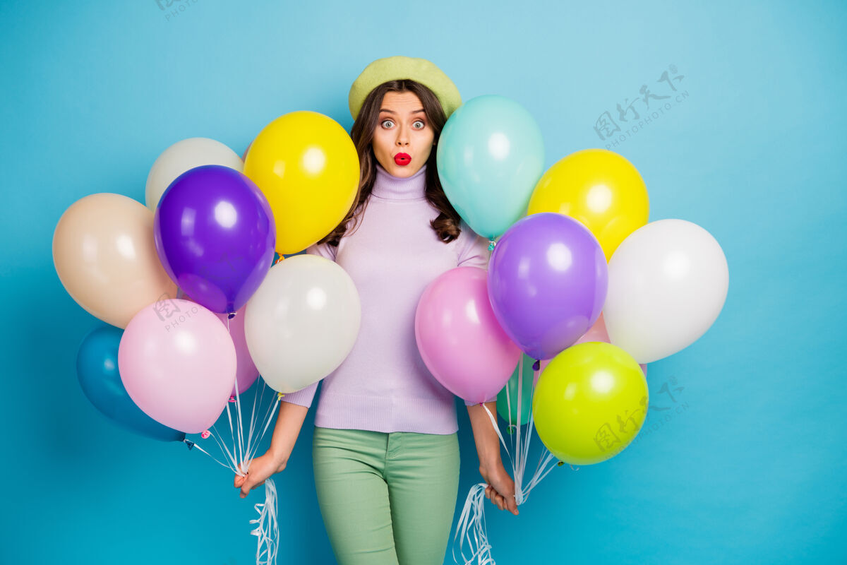 庆祝照片中搞笑的女士背着许多五颜六色的气球出人意料的惊喜派对张嘴戴紫色套头衫贝雷帽绿色裤子蓝色隔离墙冬季惊喜休闲