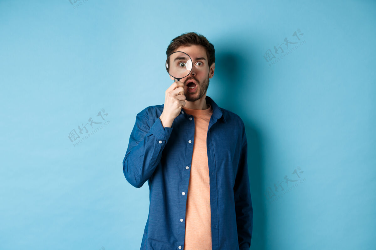 男性令人印象深刻的男人透过放大镜敬畏地看 垂下下巴盯着相机 站在蓝色的背景上表情人时尚