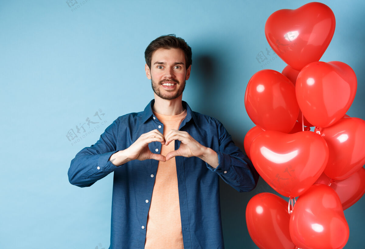 温柔情人节快乐恋爱中的男朋友向爱人展示心形 微笑着 站在蓝色背景下的红气球旁心情人情感