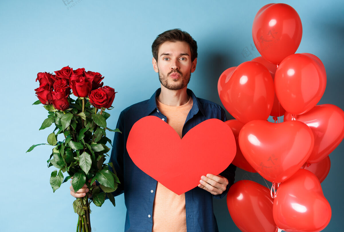 情感热恋中的男人祝福情人节快乐 在浪漫的约会中等待接吻 捧着红玫瑰 红心和气球 站在蓝色的背景上男朋友情人微笑