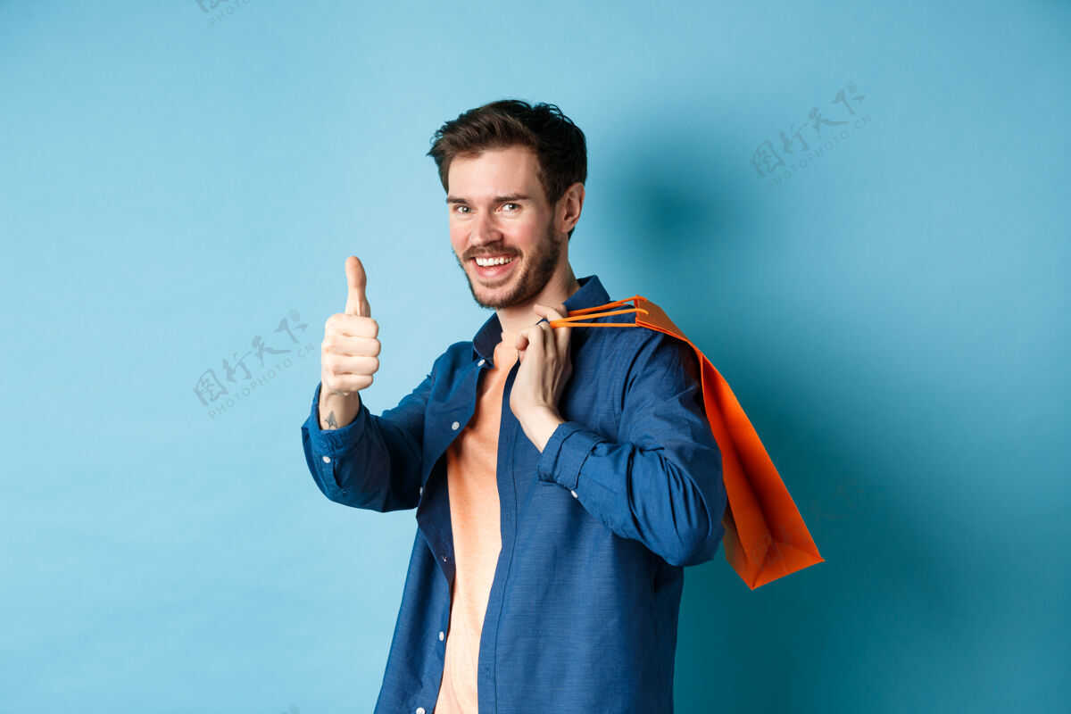 男孩帅气的年轻人 拿着购物袋 竖起大拇指 微笑着 站在蓝色的背景上 称赞好的促销优惠表情商店帅哥