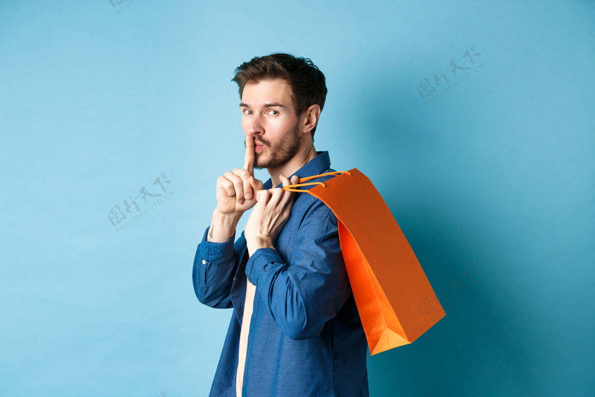 购物者现代人肩上扛着购物袋 转身对着镜头嘘嘘 讲秘密 制造惊喜 站在蓝色背景上包成人站