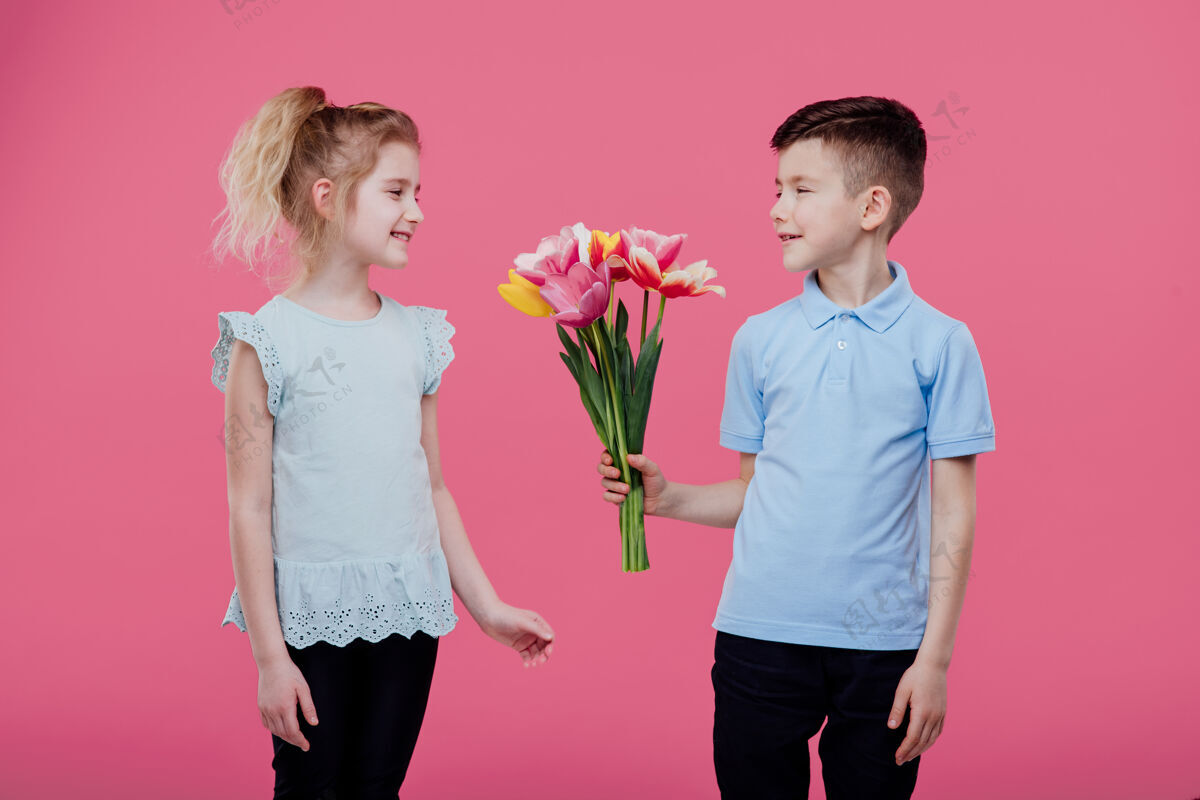 花束快乐的孩子们 男孩把花伸向一个穿着粉色裙子的小女孩 隔离在蓝色的墙上连衣裙壁板侧写