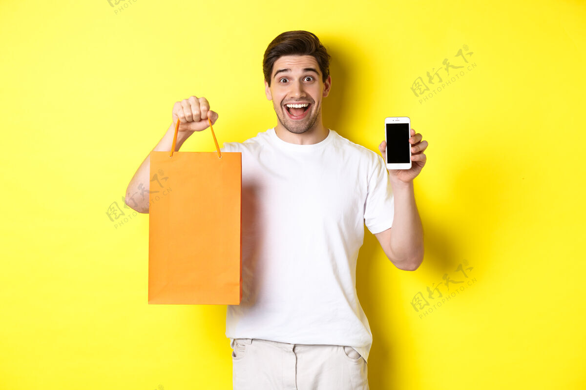应用程序惊讶的男人拿着购物袋 展示智能手机屏幕模特礼品休闲