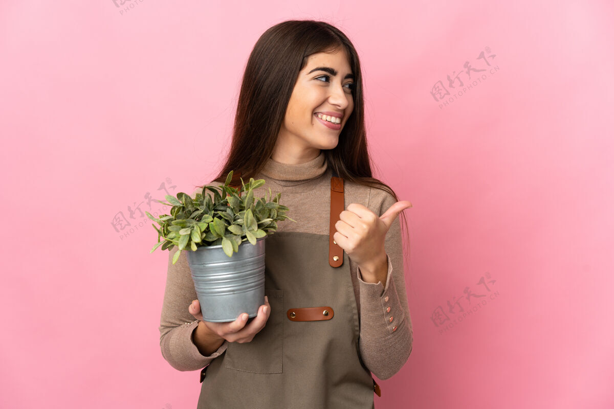 表演年轻的园丁女孩拿着一株隔离在粉红色背景上的植物 指着侧面展示产品点成人人