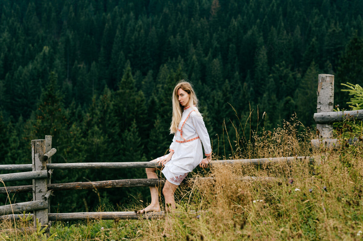 自然年轻迷人的金发女孩 穿着白色的刺绣裙 坐在风景如画的森林景观的木栅栏上乡村篱笆女人