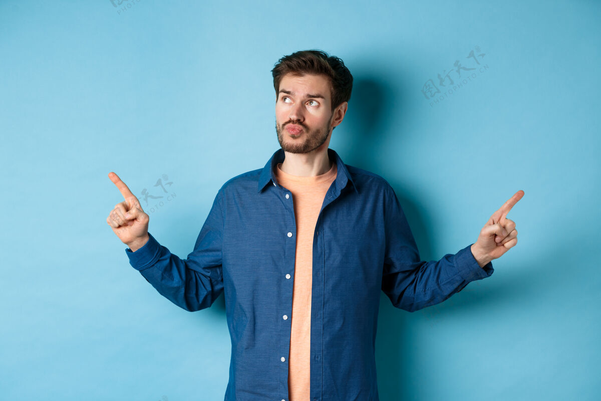 男优柔寡断的男人穿着休闲服做出选择 手指指向一边 神情沉思 站在蓝色的背景上朋友时尚肖像