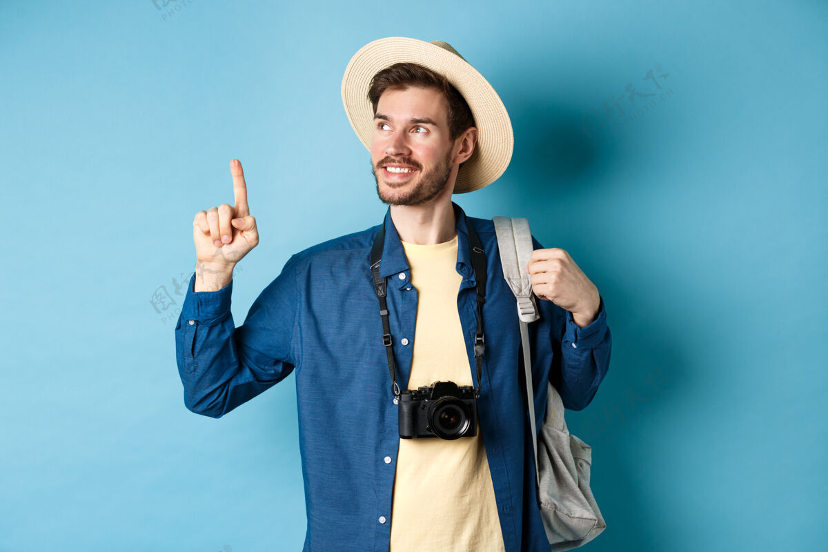 帽子度假时帅气的帅哥 戴着夏帽 抬头看着logo 高兴地笑着 拿着相机和背包站在蓝色的背景上年轻休闲指着