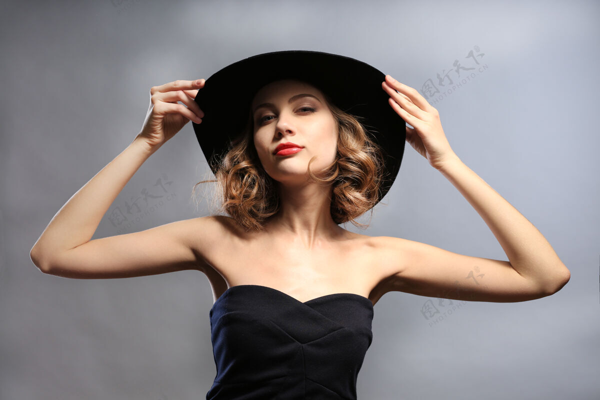 漂亮在灰色背景下 穿着黑色连衣裙戴着帽子的美丽模特的肖像年轻护理皮肤