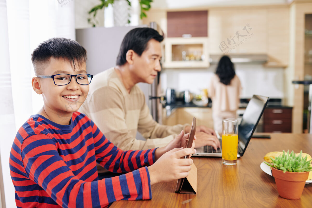 电脑一个戴着眼镜微笑的亚洲未成年男孩在餐桌旁上网学习 他的父亲在附近的笔记本电脑上工作自由职业者女人年轻人