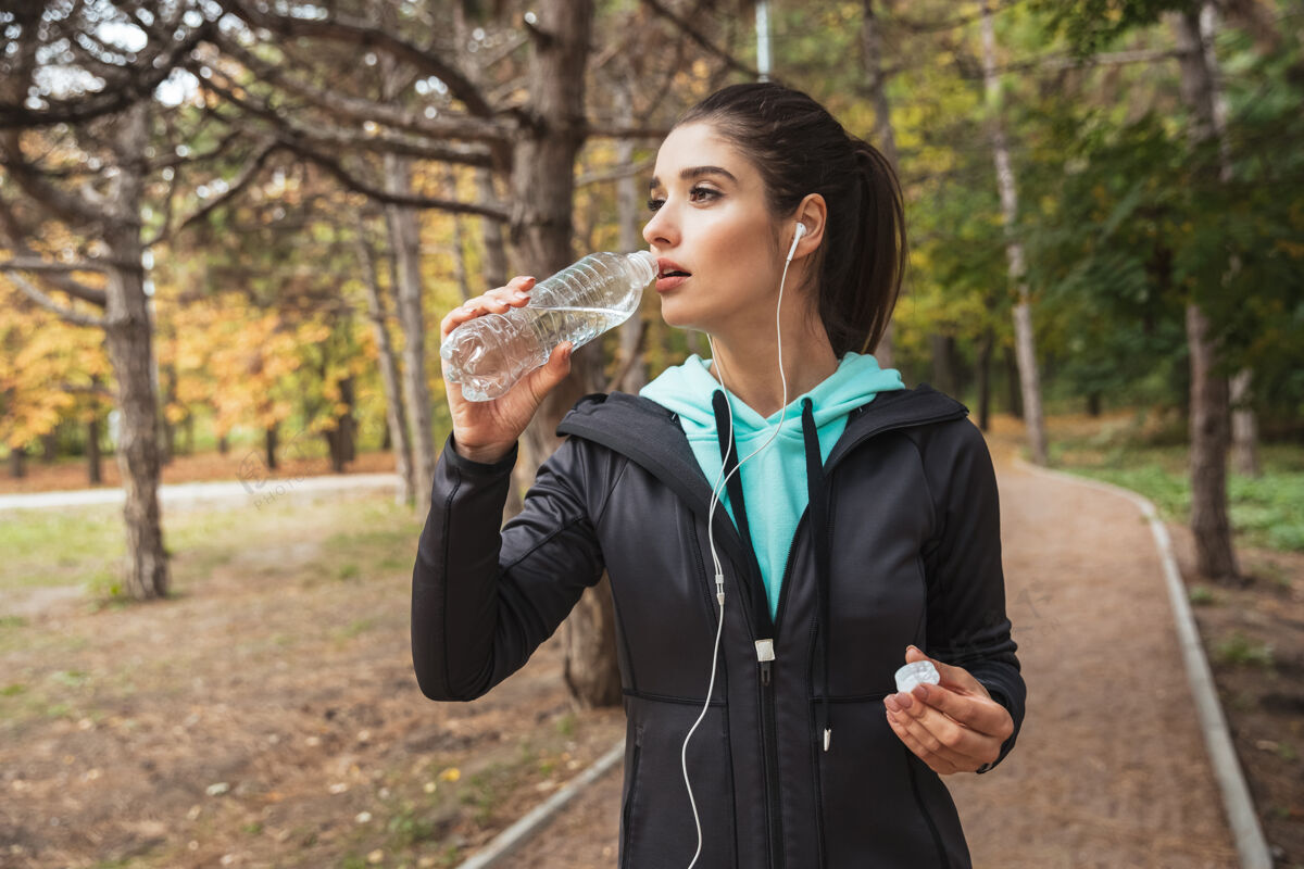 运动微笑的健身女 戴着耳机听音乐 站在公园里拿着一瓶水连接饮用水减肥