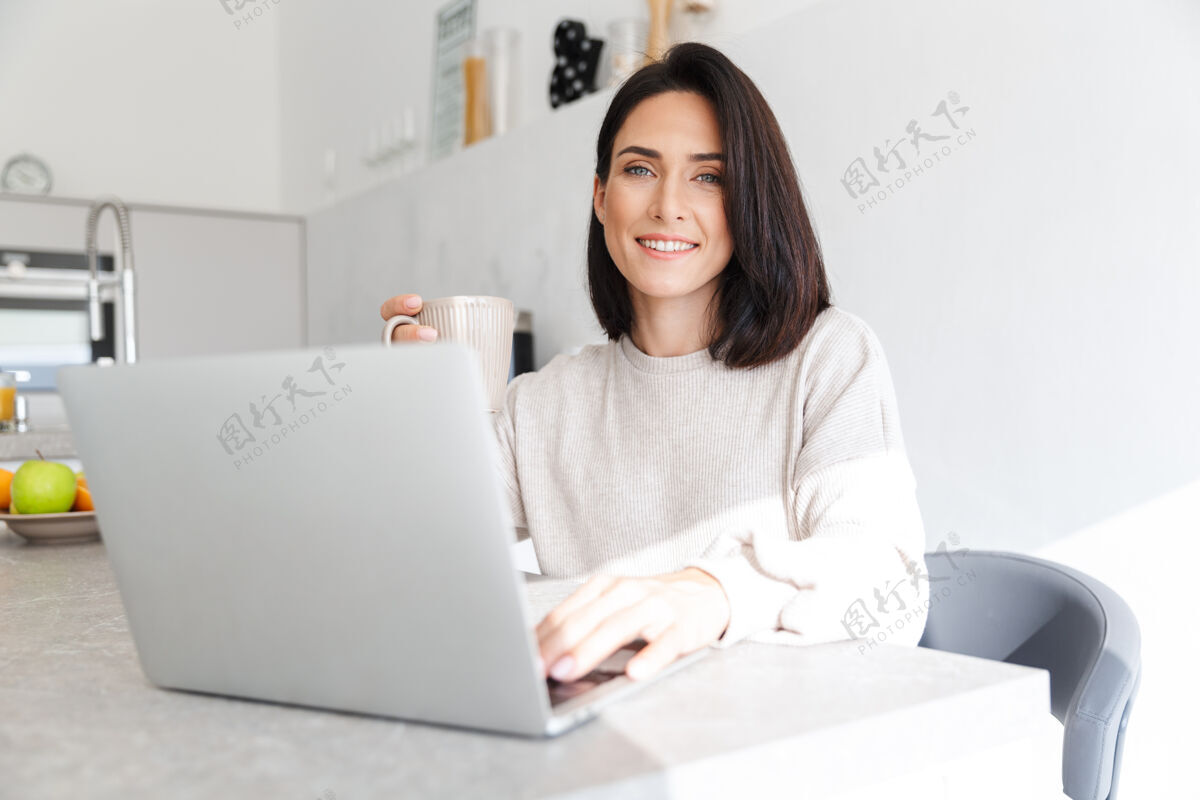 成熟30多岁的美女坐在明亮的房间里 一边用笔记本电脑 一边坐在白墙上可爱舒适笔记本电脑