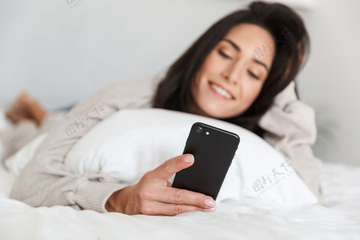 满意一张30多岁的微笑女子在明亮的房间里躺在白色亚麻布床上使用手机的照片室内成熟成人