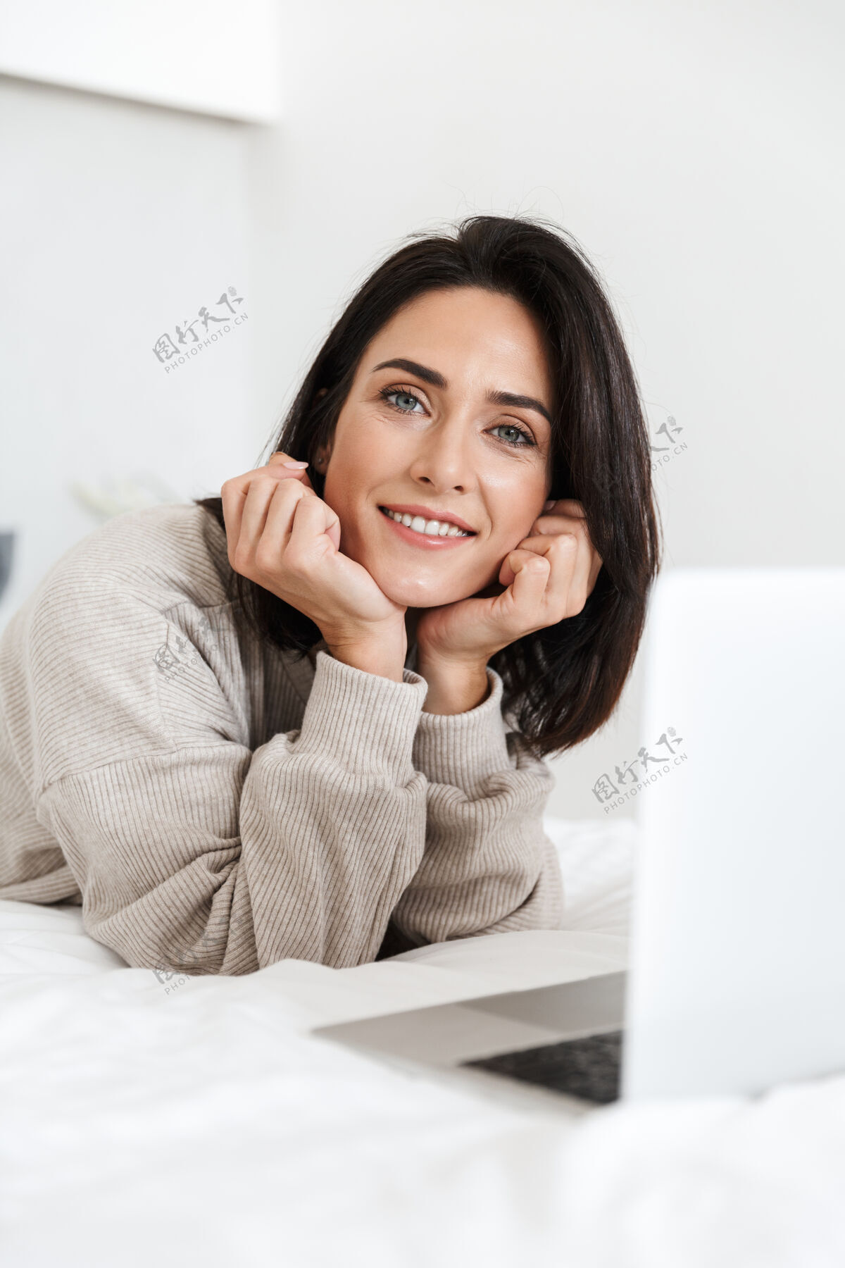 满足一张30多岁的美女在明亮的房间里躺在白色亚麻布床上使用笔记本电脑的照片深色头发放松躺着