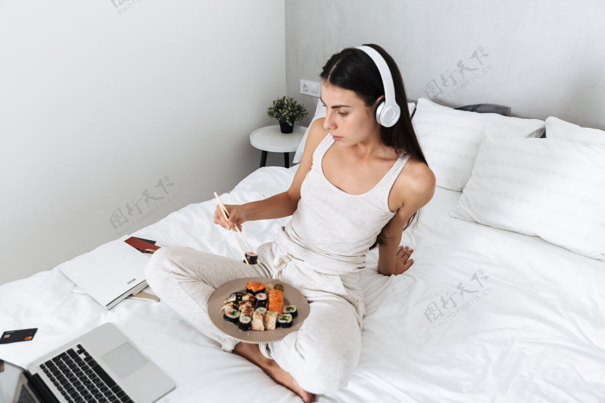 寿司美丽的年轻女子在家里的床上放松 戴着耳机听音乐 吃着盘子里的寿司阅读听力爱好