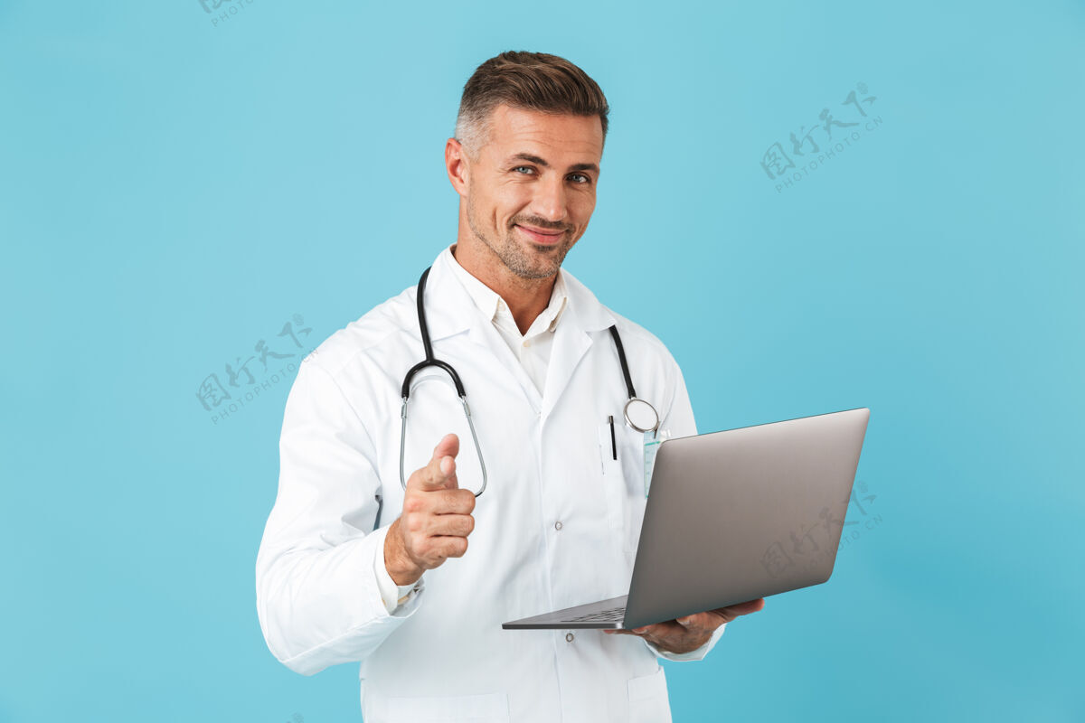 治疗照片中的帅哥身穿白色医用外套 手持手提电脑的听诊器 孤立地站在蓝色的墙上外套诊所听诊器