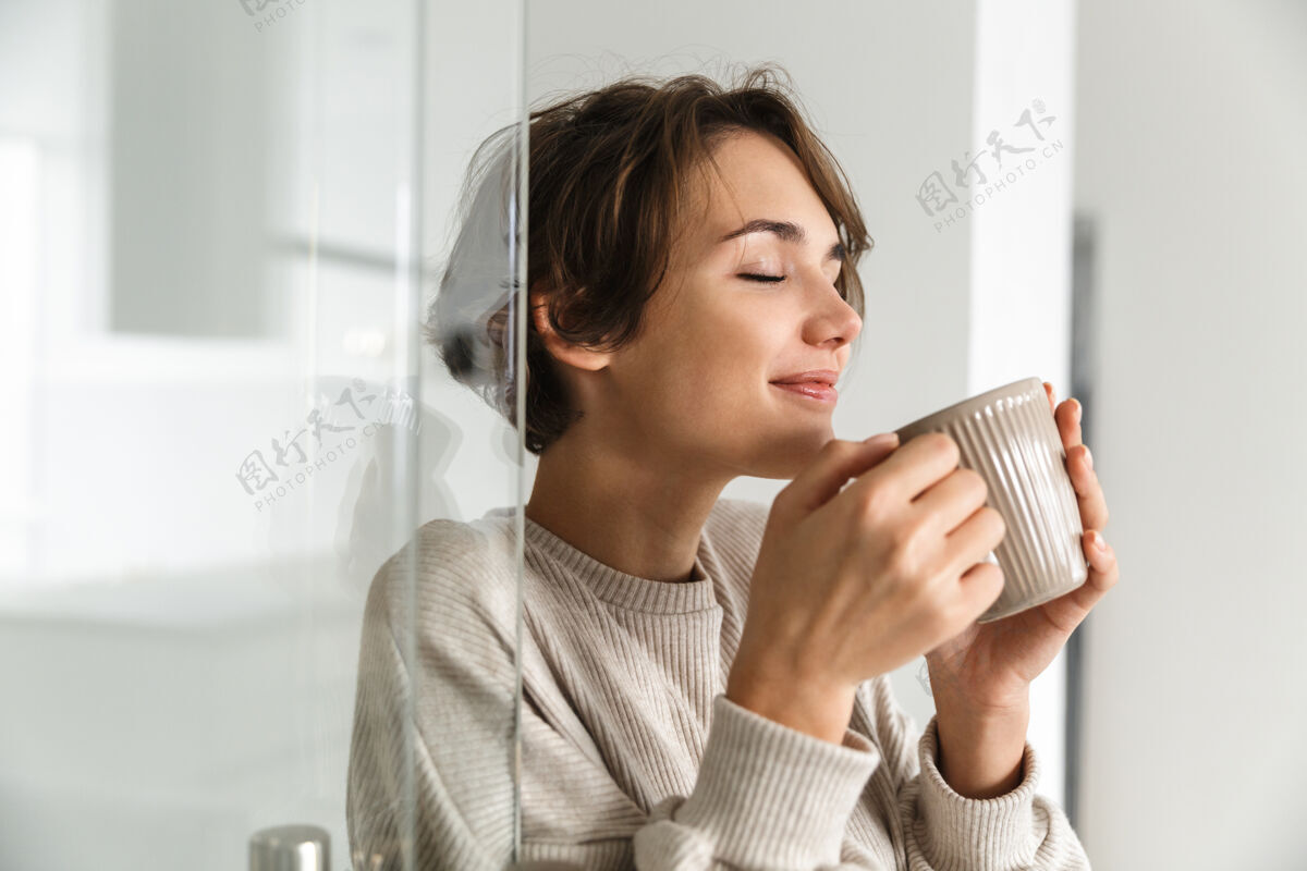 早餐快乐的黑发女人闭着眼睛喝咖啡 享受在家里室内杯子女士