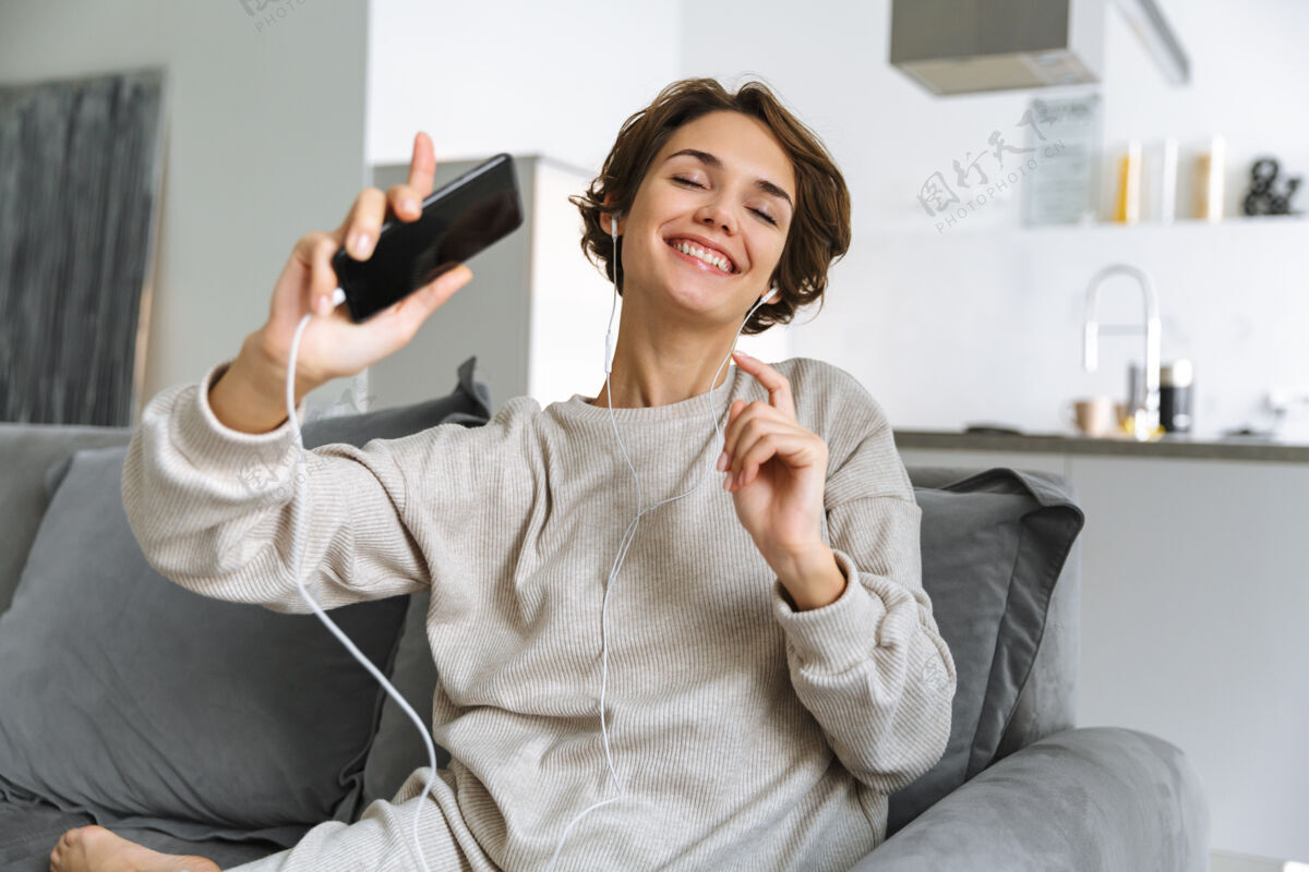 音频快乐的年轻女子坐在家里的沙发上 用手机 用耳机听音乐娱乐耳机在线
