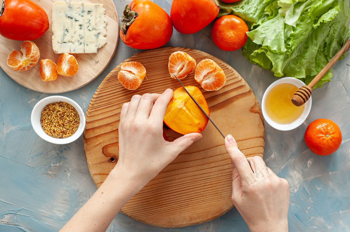盘子用柿子 橘子和蓝奶酪做维生素冬季沙拉的原料和过程一步一步地那个女人在一块圆木板上切柿子从上面看桌子美味沙拉