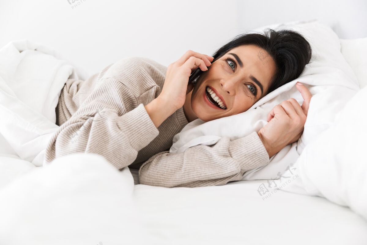 放松图为30多岁的微笑女子在家中穿着白色亚麻布躺在床上使用智能手机女人成熟手机