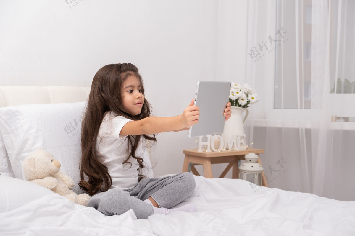 平板电脑小女孩在家里穿着泰迪熊用平板电脑坐在床上视频生活方式游戏