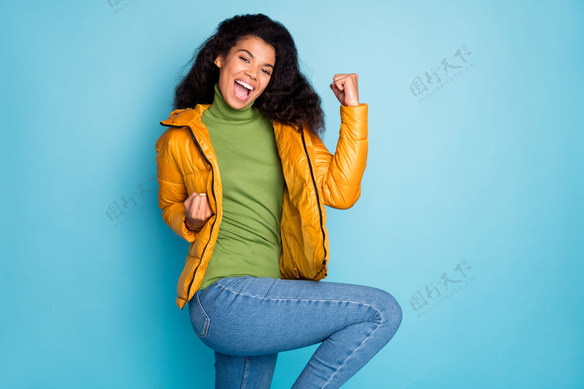 民族神奇有趣的黑色卷发女士庆祝足球比赛的目标穿时髦的黄色春季大衣牛仔裤绿色套头衫孤立的蓝色墙壁女性财富惊讶