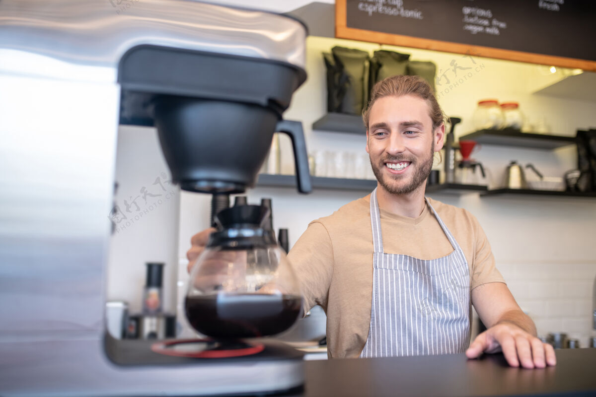 过程咖啡 微笑咖啡馆围裙里的年轻人拿着咖啡壶在咖啡馆柜台后面的咖啡机旁快乐金发胡须