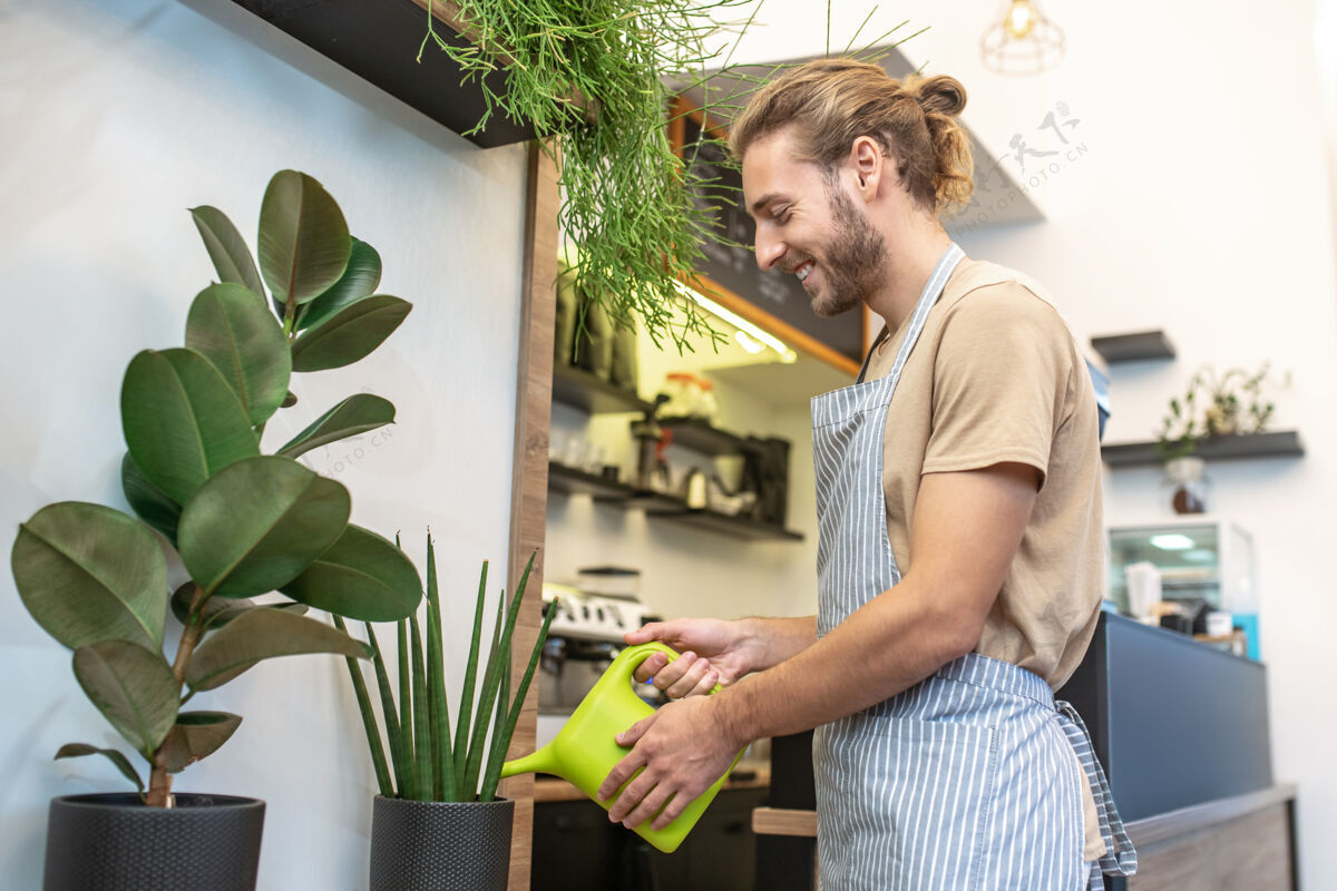男士幸运的戴微笑一个留着胡子的年轻人站在咖啡馆浇灌植物的侧面 心情很好咖啡馆工作日小生意