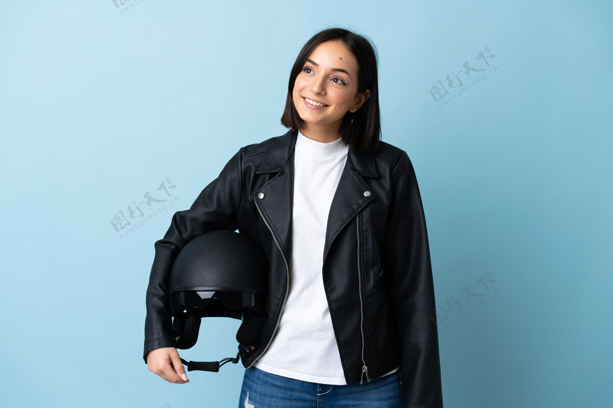 黑发一个戴着摩托车头盔的女人孤零零地站在蓝色的墙上 一边抬头一边思考着一个想法摩托车手人年轻人