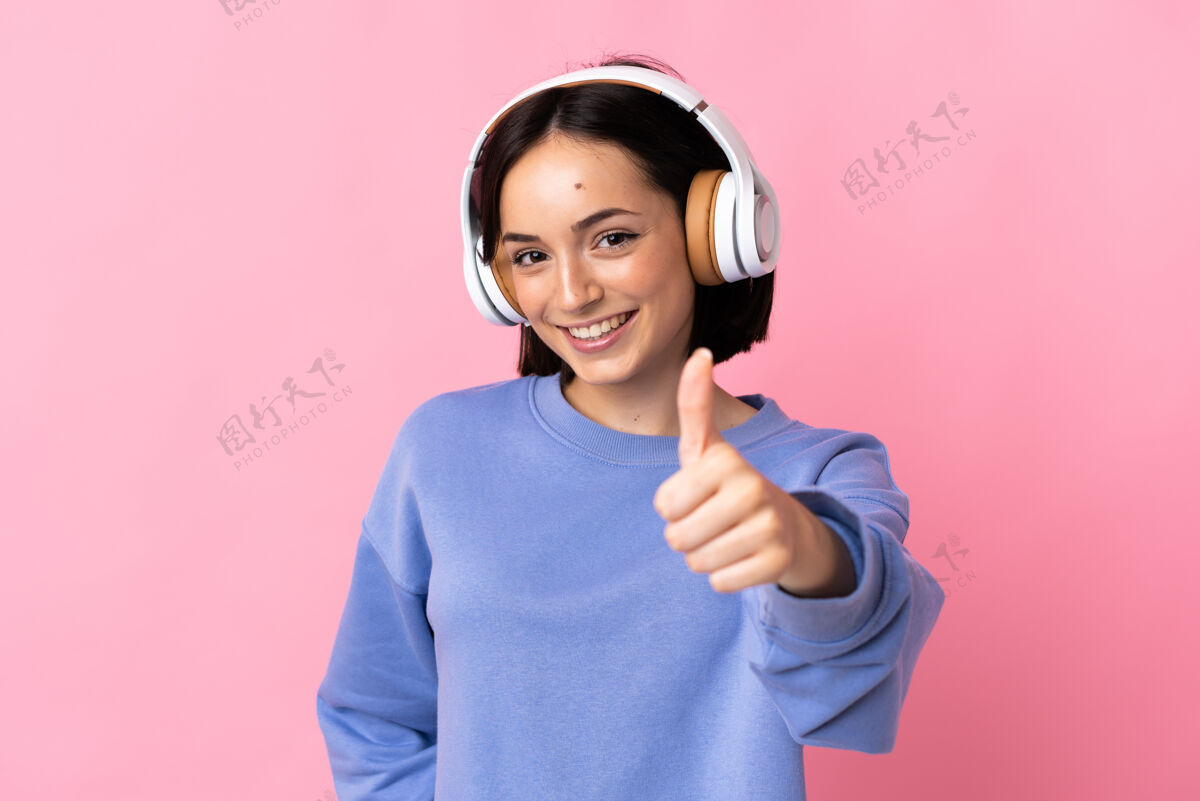 Ok一位年轻的白人妇女 孤零零地站在粉红色的墙上 一边听音乐 一边竖起大拇指女孩微笑花蕾
