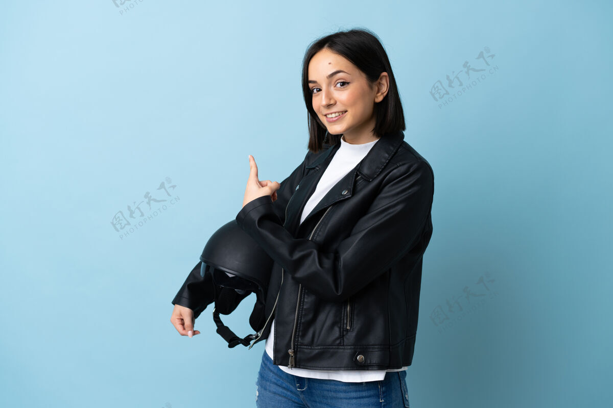 自行车一个拿着摩托车头盔的女人被隔离在蓝色的墙上指着后面手夹克女孩