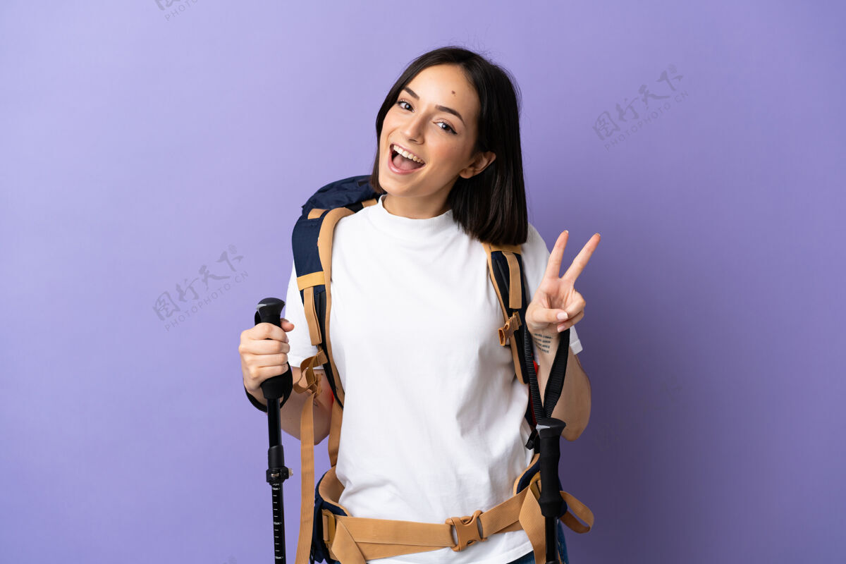 设备年轻的高加索妇女背着背包和登山竿 孤零零地站在蓝色的墙上 微笑着 展示着胜利的标志徒步旅行人和平