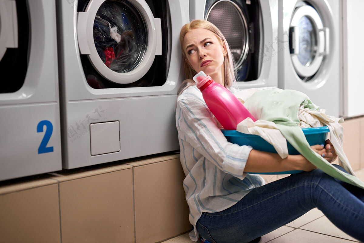 烦躁无聊的女人拿着脏衣服和洗衣粉瓶子在脸盆里 倚在洗衣机上坐在地板上 郁闷地独自坐在洗衣房里女性成人清洁