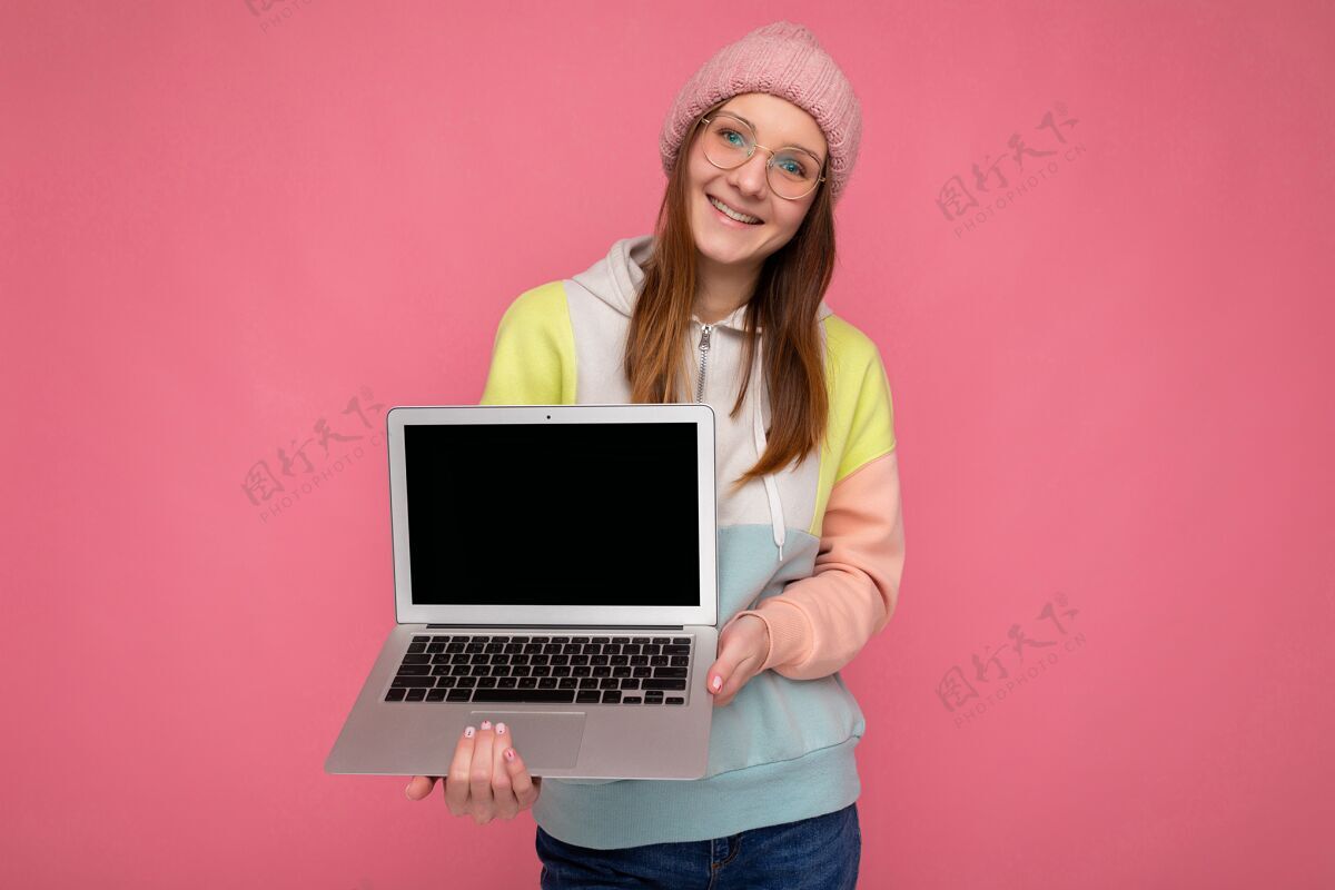 技术照片中 微笑的年轻女士戴着帽子 毛衣和眼镜 手持电脑 戴着白色耳机 看着相机背景.mock起来学习笔记本电脑上网本