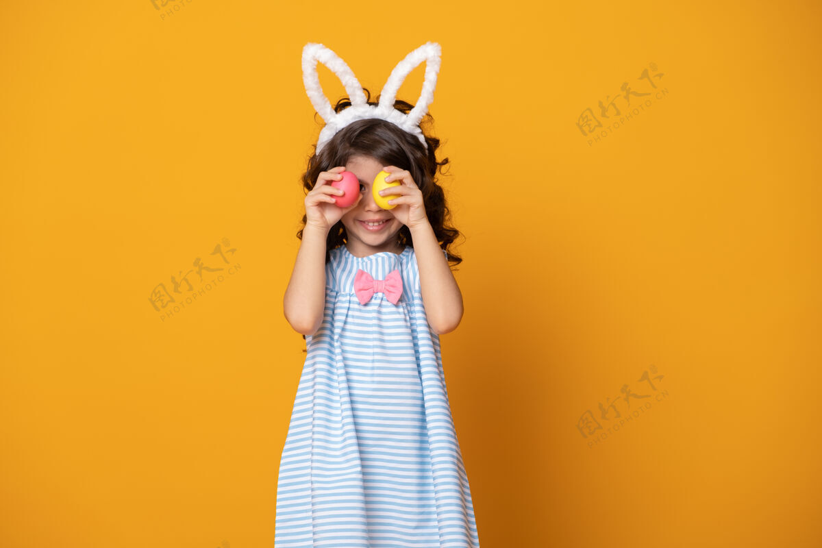 有趣有趣的小女孩戴着兔子耳朵 举着一个彩色复活节彩蛋在她眼前的黄色背景复活节微笑假日