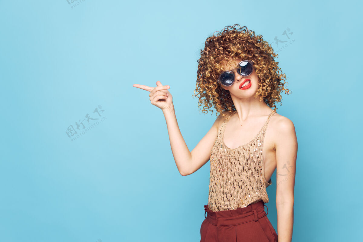 时髦女人肖像卷发用手指向侧面展示情感时尚服装特写口红微笑护目镜