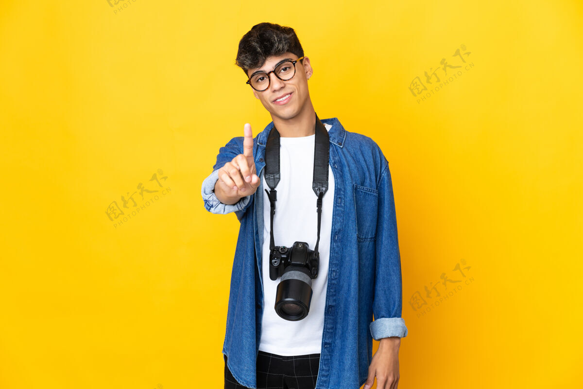 手势年轻的摄影师在隔离的黄色墙壁上展示并举起一个手指手点手指