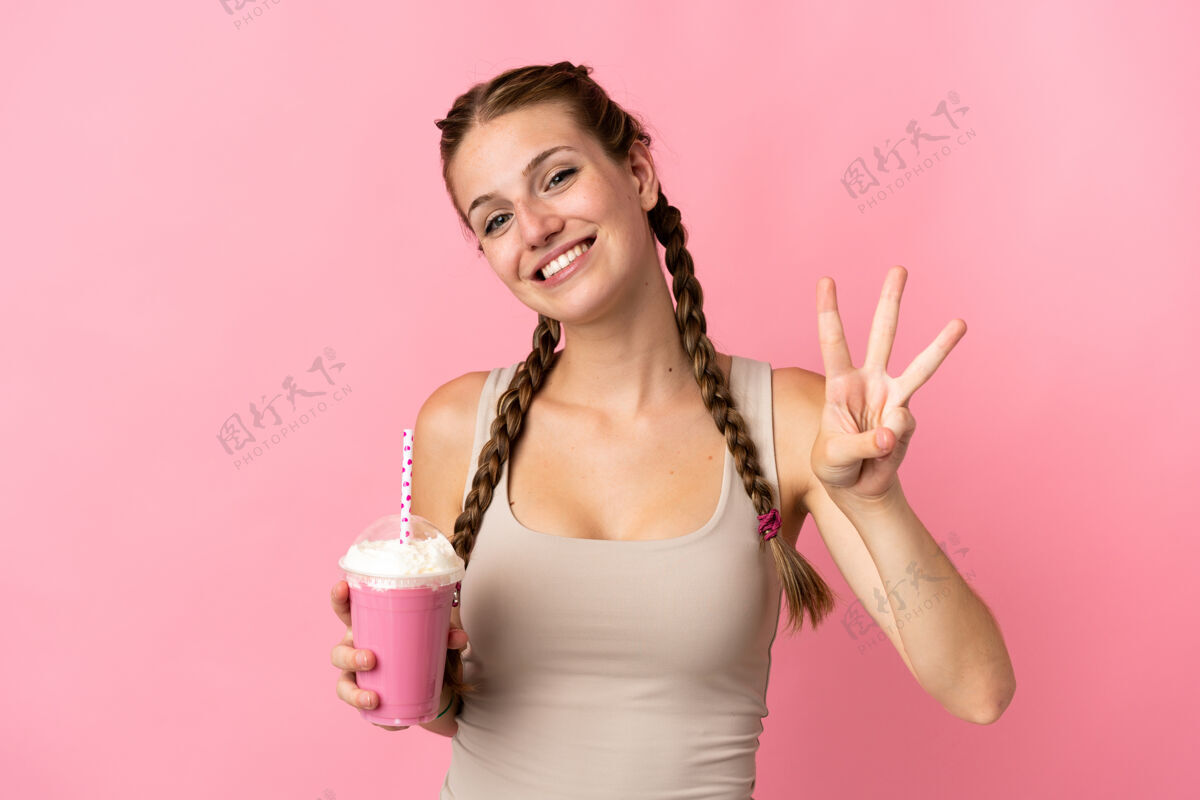 饮料拿着草莓奶昔的年轻女子在粉色背景上快乐地数着三个手指杯子甜点刷新